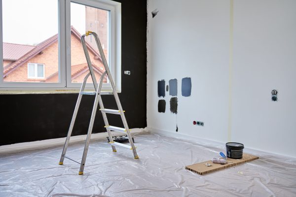 Room under renovation with color samples on white wall, ladder and painting tools on floor covered with protective film